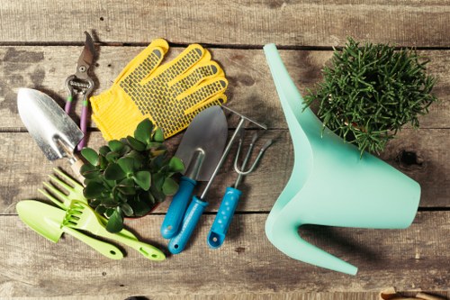 A collection of gardening tools and equipment arranged on a weathered wooden surface, including a pair of pruning shears with purple handles, a small trowel, a hand rake, a yellow gardening glove made of rubber with small ventilation holes, and a light green plant sprayer. There is a potted plant with green, needle-like foliage in a light blue watering can with a long spout positioned on the right side of the image. The background consists of the textured, natural wood surface, which provides a rustic outdoor setting typical of detailed gardening and landscaping services in Knightsbridge. The tools and plant are arranged in a balanced manner, showcasing everyday gardening tasks such as planting, pruning, and watering, emphasizing the professional outdoor maintenance and landscaping work that Landscapers Knightsbridge may undertake for clients looking to improve their garden spaces in the UK.