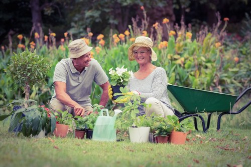 A mature couple wearing straw hats is sitting on a well-maintained lawn in a landscaped garden, surrounded by an array of potted plants and flowers. The man, dressed in a light grey polo shirt, is smiling and appears engaged in a gardening activity, while the woman, wearing a soft grey top, holds a flowering plant and is smiling warmly. Behind them, there is a lush flower bed with tall purple and orange blooms, along with green foliage creating a vibrant background. To the right, a black garden bench is positioned on the grass, and a green garden watering can is visible among the potted plants, which include a variety of flowering and leafy plants. The scene is outdoors in natural daylight, suggesting a clear, sunny day, with the garden designed for relaxation and gardening enjoyment. This image reflects typical elements of a well-kept residential outdoor space, which a professional landscaping and gardening service like Landscapers Knightsbridge might enhance with lawn care, planting, and outdoor maintenance to improve its aesthetic appeal and functionality in Knightsbridge or nearby areas.