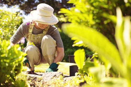 A woman working in a well-maintained residential garden, kneeling on the grass and planting or tending to flower beds bordered by stone or brick edging. She is wearing a large sun hat, gloves, and casual gardening attire. The garden features lush green foliage, including shrubs and flowering plants with vibrant and natural tones, and a neatly trimmed lawn area. In the background, there are trees providing partial shade, and the outdoor space appears to be part of a landscaped front or back garden in Knightsbridge, with natural sunlight illuminating the scene. The image showcases a peaceful outdoor environment focused on gardening activities, supporting sustainable garden practices that Landscapers Knightsbridge promote, and highlights elements like soil, plant beds, and garden borders designed for aesthetic appeal and ecological harmony.