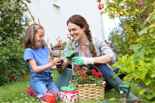 A woman and a young girl are working together in a lush, green garden likely in Knightsbridge, engaging in gardening activities during daytime under natural light. The woman, wearing a grey jacket and green gardening gloves, is smiling as she holds a small potted plant with red flowers, while the girl, dressed in a blue t-shirt and denim shorts, is assisting by holding the pot and looking at it with interest. The garden features a well-maintained lawn area in the foreground, bordered by vibrant flower beds with a variety of plants and shrubs, some with pink, red, and green foliage. A wicker basket, gardening tools, including a watering can in red and a small garden rake, are placed beside them, suggesting ongoing planting or garden maintenance. In the background, there are taller bushes and flowering plants creating a lush, natural setting. The scene captures a peaceful outdoor environment with natural tones of green, red, and earthy browns, reflecting a typical residential garden cared for by professional landscapers like Landscapers Knightsbridge, supporting local gardening and outdoor maintenance services.