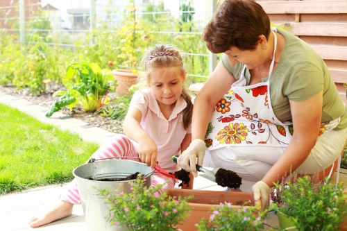 A woman and a young girl are working together in a backyard garden with a neatly maintained lawn and vibrant green plants. They are engaged in planting or tending to flower pots placed on a paved area adjacent to a wooden shed. The woman is wearing a floral apron and light-colored gloves, leaning over to assist the girl, who has long light brown hair and is dressed in a light pink top and striped leggings. The garden background features a wire fence, with various bushes and flowering plants, and potted plants on the ground. The scene is illuminated by natural daylight, with a bright, clear outdoor environment that suggests mild weather. This outdoor space demonstrates typical landscaped garden features with flower beds and potted plants, and visually aligns with professional gardening services offered by companies such as Landscapers Knightsbridge to enhance outdoor spaces in areas like Knightsbridge or surrounding locations.