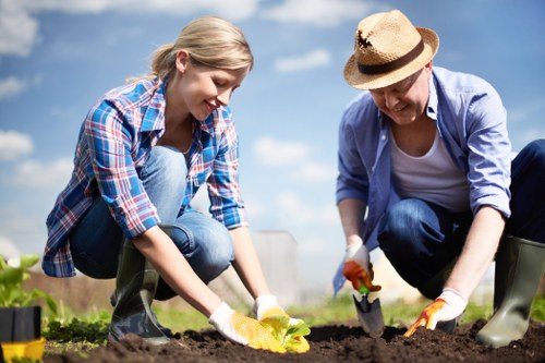 A young woman and an older woman are engaged in planting activities in a well-maintained outdoor garden, with the younger woman wearing a blue plaid shirt and jeans, and the older woman dressed in a light blue shirt, jeans, and a straw hat. They are kneeling on the soil, using small garden trowels to dig and plant, indicating ongoing gardening work. The garden features rich, dark soil with visible tilled bed areas, and there are gardening tools and potted plants nearby. The background includes a bright blue sky with some clouds, suggesting a clear, sunny day, ideal for outdoor gardening in the area near Knightsbridge. This scene is representative of professional gardening services offered by Landscapers Knightsbridge, suited for residential or landscaped outdoor spaces in the local area.