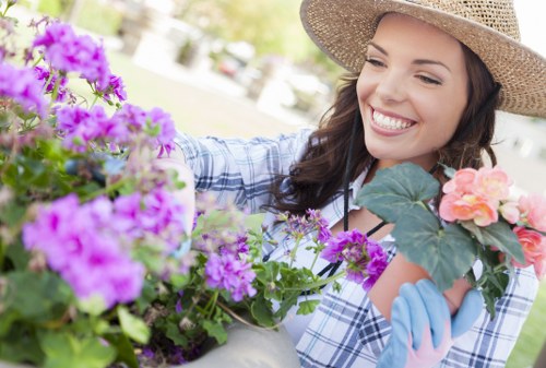A smiling woman in a straw hat and light blue gardening gloves tends to vibrant flowering plants in a landscaped garden. The garden features a variety of plants, including purple and pink flowers, with dense green foliage and well-maintained flower beds. In the background, a neatly trimmed hedge and trees are visible, set in a sunny outdoor environment with natural daylight illuminating the scene. The garden appears organized and designed for aesthetic appeal, likely part of a private yard or communal outdoor space. The woman’s focus on the plants suggests ongoing gardening or maintenance activity, aligned with professional gardening services offered by Landscapers Knightsbridge. The scene captures the natural textures of soil, plant leaves, and blossoms, emphasizing quality outdoor care and horticultural attention, typical of high-standard landscaping in the London area, including Knightsbridge postcode.