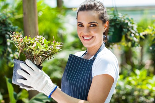 A smiling woman with dark hair tied back, wearing a navy blue striped apron and white gardening gloves, is standing in a lush garden during daytime. She holds a small potted shrub with green and pink-tinted leaves, positioned slightly to the left of her. The background features vibrant greenery with various plants, including tall trees and flowering bushes, suggesting a well-maintained outdoor space. The scene appears to be in a garden or backyard, with natural sunlight illuminating her face and the surrounding foliage, creating a bright and inviting atmosphere that supports gardening and landscaping services in the Knightsbridge area. The environment is characterized by healthy, dense plant life and a clean outdoor setting, emphasizing horticultural care and garden maintenance.