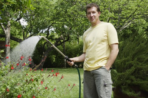 A man in a light yellow T-shirt and grey cargo shorts stands in a well-maintained garden, holding a garden hose and watering a bed of red flowering plants. The garden features a lush green lawn in the foreground, bordered by flowering plants, with mature trees and dense shrubbery in the background, providing a natural, landscaped outdoor space. The scene is set on a bright, partly cloudy day, with dappled sunlight filtering through the tree canopy, highlighting the vibrant colors of the flowers and the healthy green foliage. The garden is part of a landscaped yard typical of residential properties serviced by companies like Landscapers Knightsbridge, focusing on outdoor irrigation, lawn care, and planting management within the Knightsbridge area. The environment appears tidy and professionally maintained, with the garden hose indicating ongoing outdoor maintenance or watering tasks, aligning with expert gardening and landscaping services in the local area.