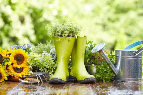 A wooden outdoor table in a garden setting showing a pair of bright green rain boots filled with small white and green flowering plants, surrounded by gardening tools and flower arrangements. To the left, there are vibrant yellow sunflowers with dark centers and purple flowers, while on the right, a metal watering can with a blue handle is positioned beside lush green foliage. The background features a softly blurred garden scene with greenery and sunlight filtering through trees, indicating a well-maintained outdoor space typical of a garden in Knightsbridge. The surface of the table appears slightly wet, suggesting recent watering or rain, and the scene reflects careful outdoor garden care and landscaping efforts consistent with professional gardening services offered by Landscapers Knightsbridge.