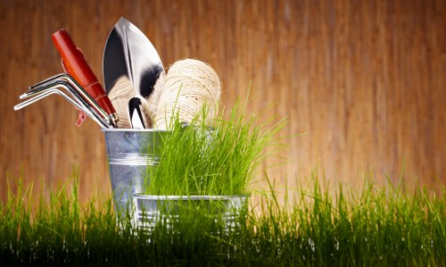 The image shows a close-up view of two metal gardening containers placed on a neatly maintained grassy area, likely in an outdoor garden space in Knightsbridge. The containers include a small bucket holding gardening tools such as a hand rake, a trowel, and a gardening fork, alongside a ball of twine and a bunch of vibrant green grass-like plants. The background features a warm, wooden surface that provides a natural ambiance, complementing the outdoor setting. The lush, dense grass in the foreground frames the containers, emphasizing the tools and planting materials prepared for garden maintenance or creation. The scene appears to be set on a bright, clear day, with natural light highlighting the textures of the grass, the metallic sheen of the containers, and the wooden backdrop, illustrating a well-organized outdoor gardening environment suitable for a professional landscaping service in Knightsbridge or surrounding areas.