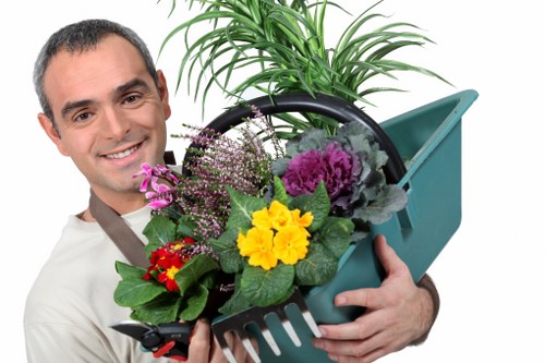 The image shows a smiling man holding a teal plastic garden tote filled with a variety of vibrant flowering plants and green foliage. The plants include purple and pink blossoms, a bright yellow flower, and broad green leaves, all arranged together in the tote. Behind him, there is a lush green potted plant with long, arching leaves, indicating a healthy, well-maintained garden environment. The outdoor setting suggests a landscaped area, possibly part of a residential garden in Knightsbridge, with the natural light highlighting the lively colours of the flowers and the greenery. The overall scene emphasizes professional gardening and plant care, aligning with services offered by landscaping specialists such as Landscapers Knightsbridge, who focus on garden maintenance, planting, and outdoor aesthetics. The image communicates a professional, approachable gardening expert prepared to enhance outdoor spaces with vibrant plant selections and attentive care, suitable for garden projects across London and nearby areas including postcode SW1X.