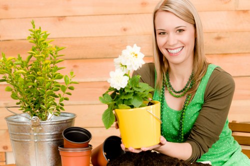 The image shows a woman with blonde hair and a cheerful expression, sitting outdoors in a garden area with a wooden fence in the background. She is holding a yellow ceramic pot containing a flowering plant with white blooms and green foliage. In front of her are several empty terracotta and black plastic plant pots, indicating gardening activities. To her left, there is a large potted shrub or small tree in a metallic container. The garden surface appears to be soil or mulch, with additional potted plants nearby. The overall scene suggests a peaceful outdoor space for gardening and plant care, aligning with landscaping services like planting, potting, and garden maintenance, which are typical of companies such as Landscapers Knightsbridge. The natural lighting and weather conditions imply a clear day suitable for garden work, highlighting the importance of sustainable planting and garden sustainability practices in London and nearby areas.