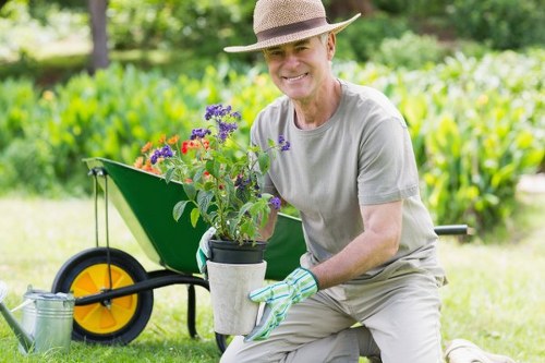 A smiling older man wearing a straw hat, light grey t-shirt, and gardening gloves kneels on a well-maintained lawn in a garden. He is holding a small potted plant with colorful purple and red flowers, which he has just lifted from a green wheelbarrow visible in the background. The garden features lush green grass, neatly trimmed hedges, and mature trees, with sunlight filtering through the foliage, creating a bright outdoor environment. The scene is indicative of gardening or landscaping activities in a residential garden setting, with natural tones of green, soil, and vibrant plant colours. This setting aligns with the services offered by Landscapers Knightsbridge, focusing on outdoor gardening care and landscape enhancement around Knightsbridge, SW1.