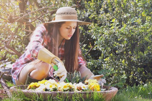 A woman wearing a wide-brimmed straw hat, gardening gloves, and casual clothing is crouched down in a lush garden, tending to a circular flower bed with yellow, white, and purple flowers. Around her, well-maintained green grass and dense shrubbery form a natural outdoor environment, indicating a landscaped garden in Knightsbridge. The scene is illuminated by natural sunlight, suggesting a clear day, with background foliage providing a vibrant, natural backdrop. The structured flower bed contrasts with the surrounding grassy area, demonstrating professional landscaping practices, and the image reflects outdoor gardening activities typical of quality garden maintenance and landscaping services in the Knightsbridge area.