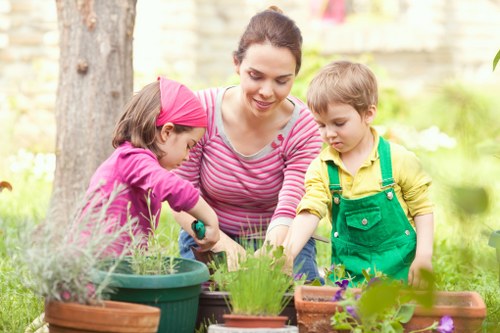 A woman and two young children are engaged in gardening activities in an outdoor garden area. The woman, dressed in a pink and white striped top, is smiling as she assists the children in tending to various potted plants. One child, wearing a pink cap and pink jacket, is actively watering or handling a plant in a green pot, while the other child, dressed in a yellow shirt and green apron, is observing or participating nearby. The garden features a well-maintained grassy lawn, several flower pots with different plants, some with purple flowers and lush green foliage. In the background, there are trees with thick trunks and a blurred garden fence, indicating a secure and private outdoor space. The scene is set during daylight with natural sunlight illuminating the scene, reflecting a pleasant and nurturing environment ideal for family gardening and outdoor activities, supported by professional landscaping services such as those provided by Landscapers Knightsbridge near the Knightsbridge area in London, England.