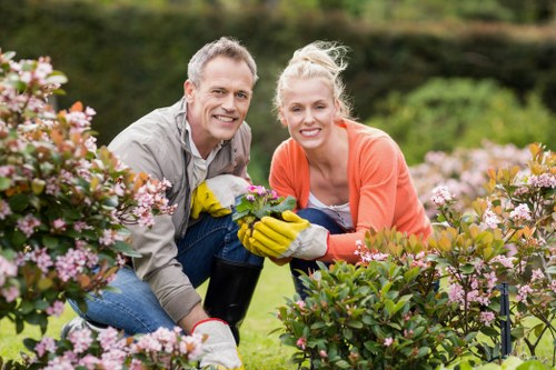 A cheerful man and woman in a well-maintained garden, both wearing gardening gloves and casual work clothes, are kneeling amidst vibrant, pink-flowering bushes. They are smiling at the camera while tending to the bushes, with one of them holding a watering can. The garden features neatly arranged shrubs, a lush green lawn, and a backdrop of outdoor trees and open space, suggesting a landscaped backyard or front garden typical of homes in the Knightsbridge area. The scene is set in bright natural daylight, indicating clear weather, and highlights the company's gardening services, including shrub and flower bed maintenance. The overall environment emphasizes a healthy, vibrant outdoor setting suitable for local gardening and landscaping tasks.